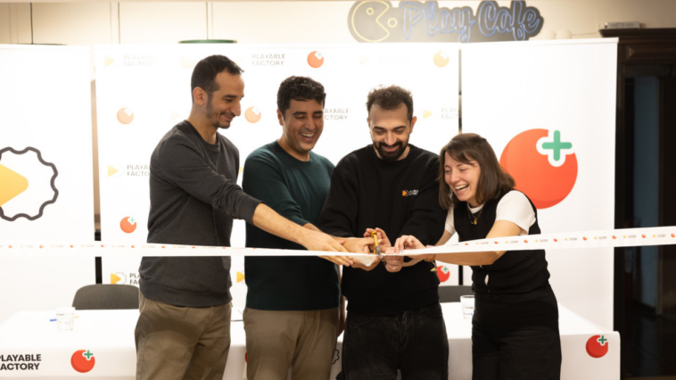 Four people cut a celebratory ribbon together, smiling, at an event backdrop with logos and a Playable Factory sign in the background.